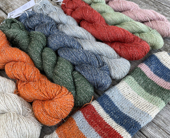 8 skeins of different coloured yarns are laying on a wooden table with a long swatch, using all the colours, in front.