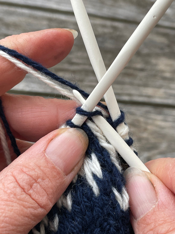 a pair of hands holding double pointed knitting needles whilest knitting with 2 a white and a blue strand creating some colour work.
