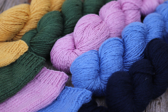 Image shows 5 skeins of yarns and swatches in different colours: from right to left navy blue, a sky blue, pink like cheery blossoms, British racing car green and mustard yellow yellow, 